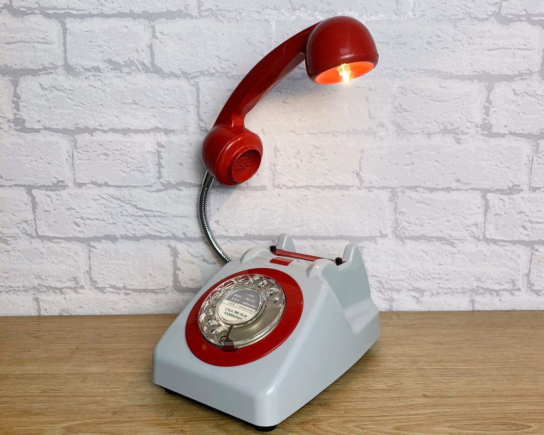 A quirky lamp made from a retro phone. The base of the lamp is a classic pale grey rotary phone with a red-colored dial ring. The red handset mounted on a flexible gooseneck has a small LED bulb in the receiver converting it into a fabulous lamp.