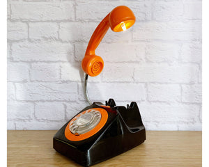 A quirky lamp made from a retro phone. The base of the lamp is a classic black rotary phone with a bright orange dial ring. The orange handset mounted on a flexible gooseneck has a small LED bulb in the receiver converting it into a fabulous lamp.