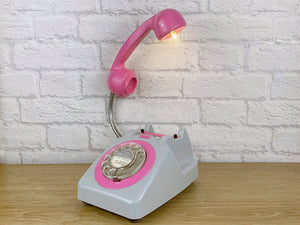 A quirky lamp made from a retro phone. The base of the lamp is a light grey rotary phone with a hot pink dial ring. The hot pink handset mounted on a flexible gooseneck has a small LED bulb in the receiver converting it into a fabulous lamp.